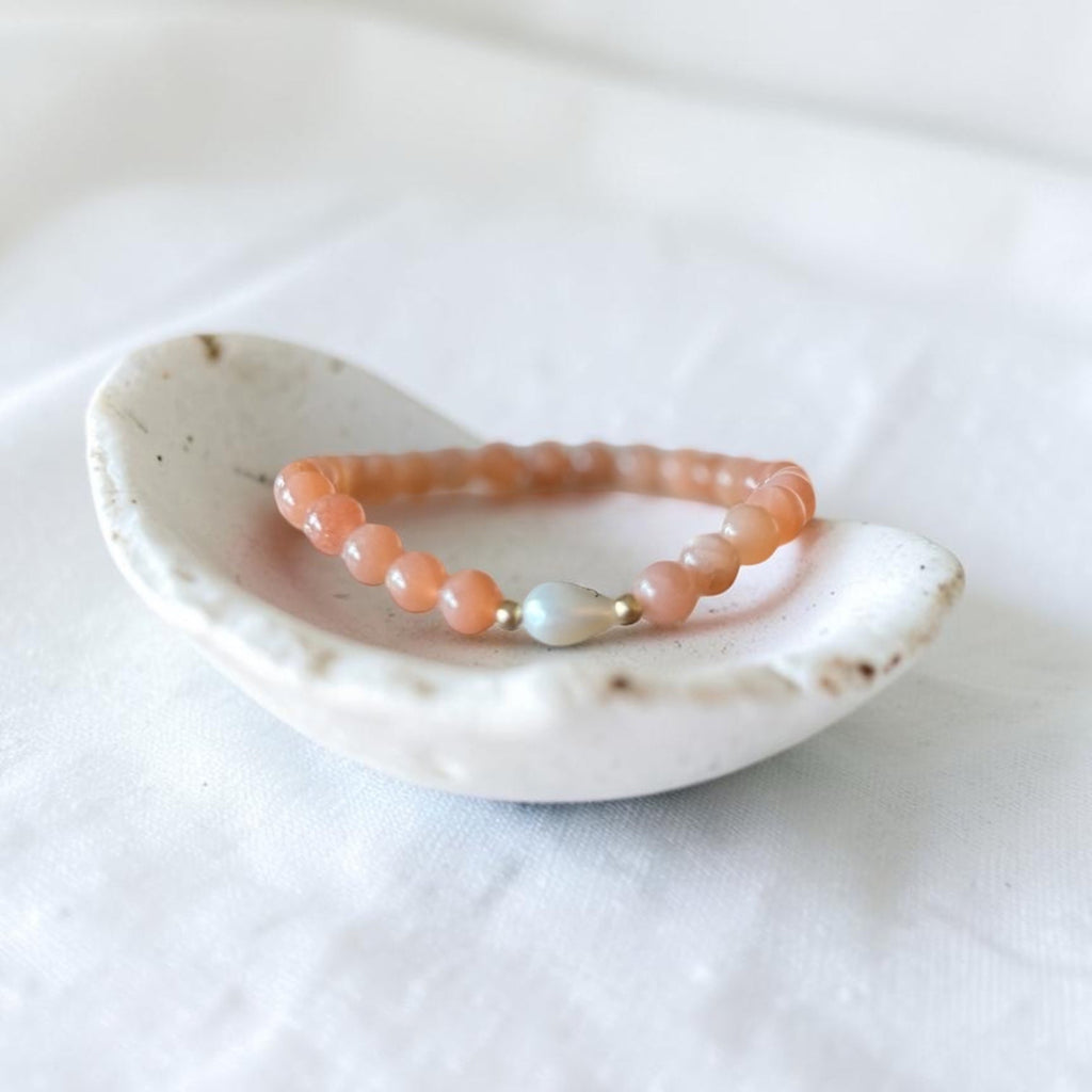 A beaded stretch bracelet in peachy orange beads with a single white pearl-like bead and a tiny gold spacer, displayed on a small white ceramic dish over a white fabric background.