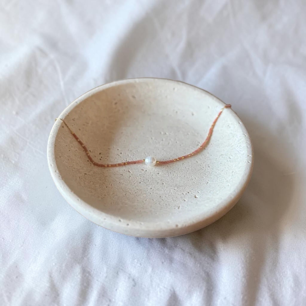 A delicate brown cord necklace with a single small white pearl bead, displayed inside a small, round, speckled beige ceramic dish on a white fabric background.