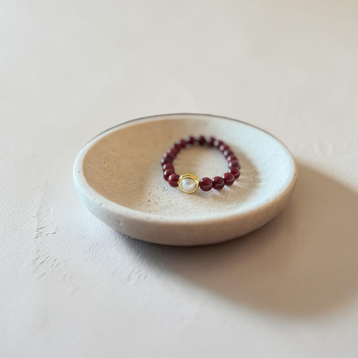8mm Red Garnet Jade Bead Bracelet with a Gold And Pearl Accent. Accent has a round pearl with gold surrounding the pearl itself. This bracelet is sitting in a dish.