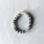 A beaded stretch bracelet featuring gray and white speckled stone beads, white faux pearls, and small gold spacer beads, displayed on a textured off-white background.