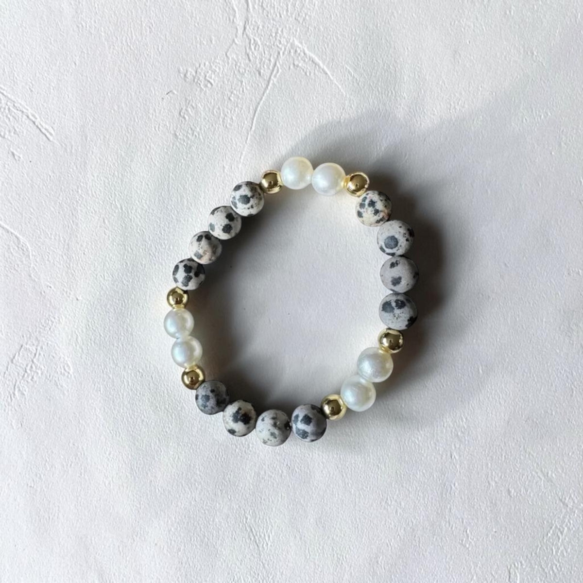A beaded stretch bracelet featuring gray and white speckled stone beads, white faux pearls, and small gold spacer beads, displayed on a textured off-white background.