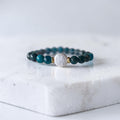 A beaded stretch bracelet featuring dark teal and black beads, a central textured white round bead, and small gold spacer rings, displayed on a white marble block.