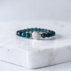 A beaded stretch bracelet featuring dark teal and black beads, a central textured white round bead, and small gold spacer rings, displayed on a white marble block.