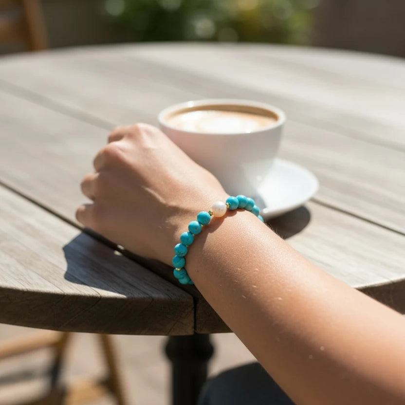 A turquoise beaded bracelet with a small white bead accent worn on a person's wrist, shown resting on a wooden table next to a cup of coffee in an outdoor setting.