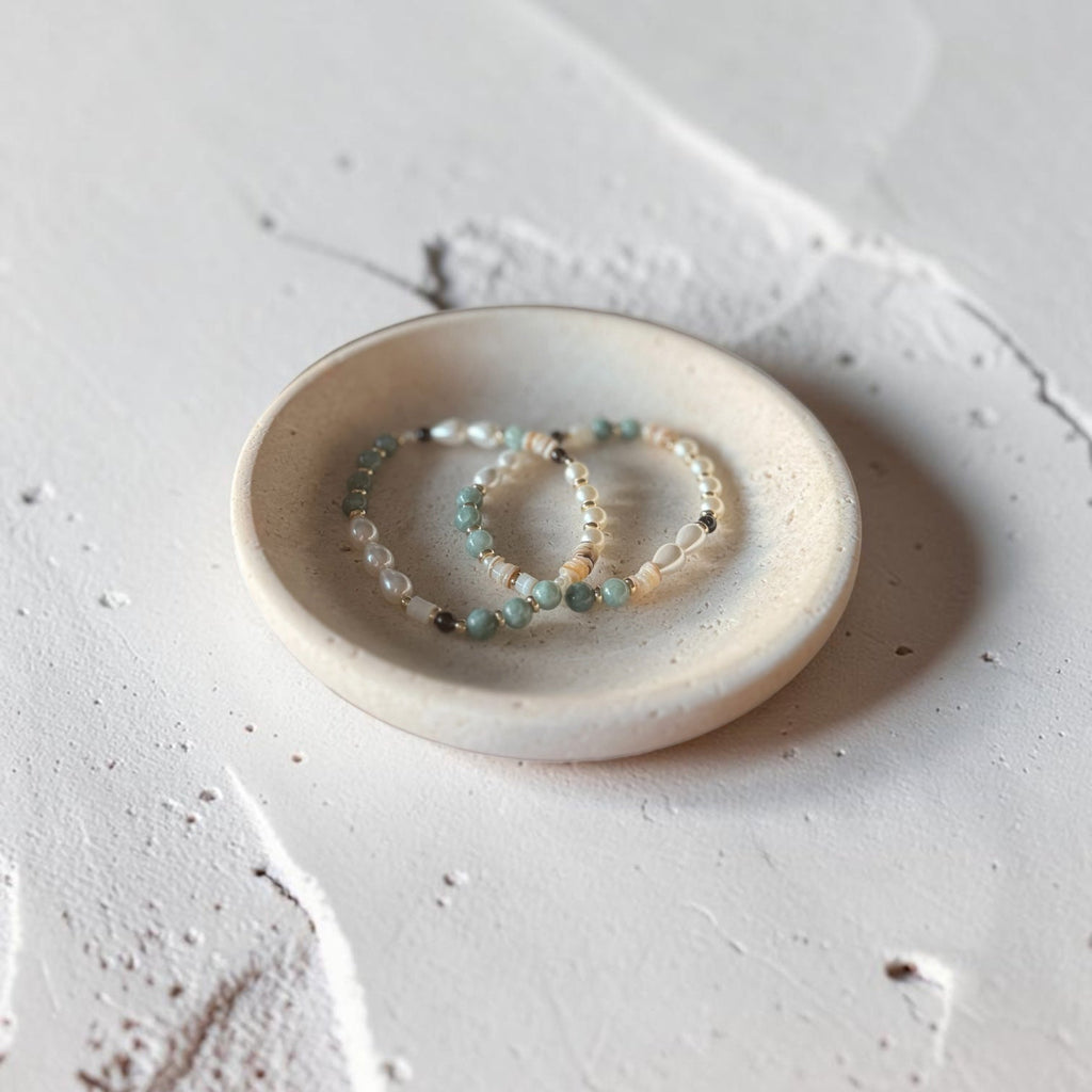 Two beaded bracelets with green, white, and dark gray beads resting on a small round ceramic dish, set against a textured light gray concrete background.