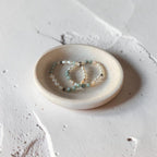 Two beaded bracelets with green, white, and dark gray beads resting on a small round ceramic dish, set against a textured light gray concrete background.