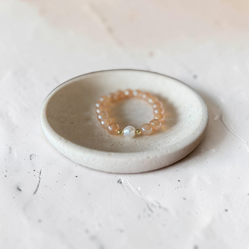 A beaded stretch bracelet with peach-orange translucent beads, a white pearl-like bead, and small gold spacer beads, displayed on a small round ceramic dish over a textured light surface.