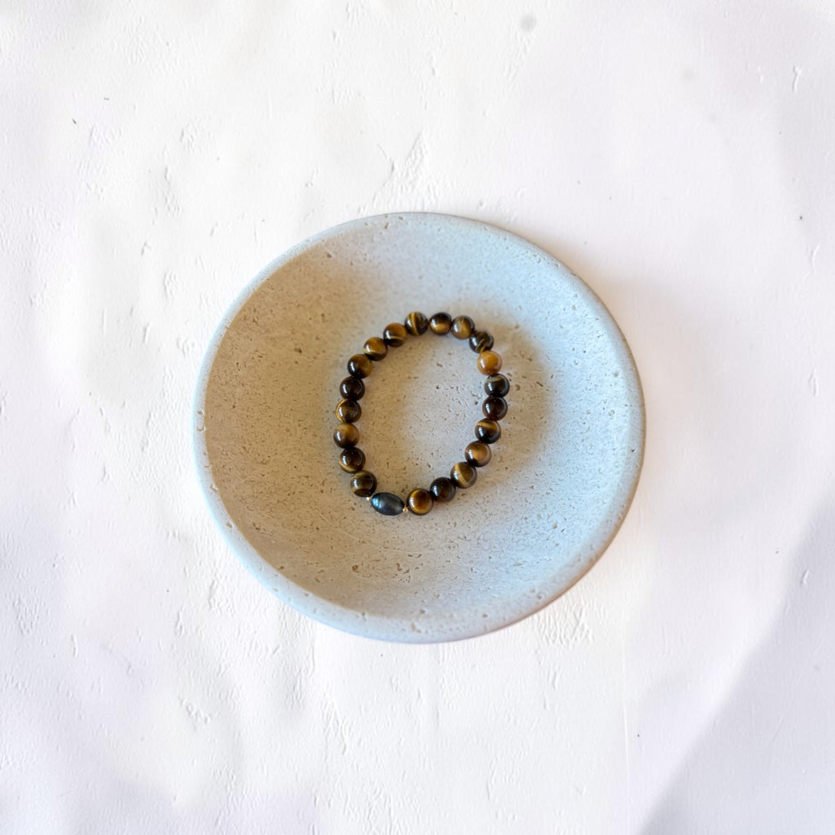 A beaded stretch bracelet made of dark brown and black tiger's eye beads, displayed inside a small, round, speckled ceramic dish on a textured white surface.