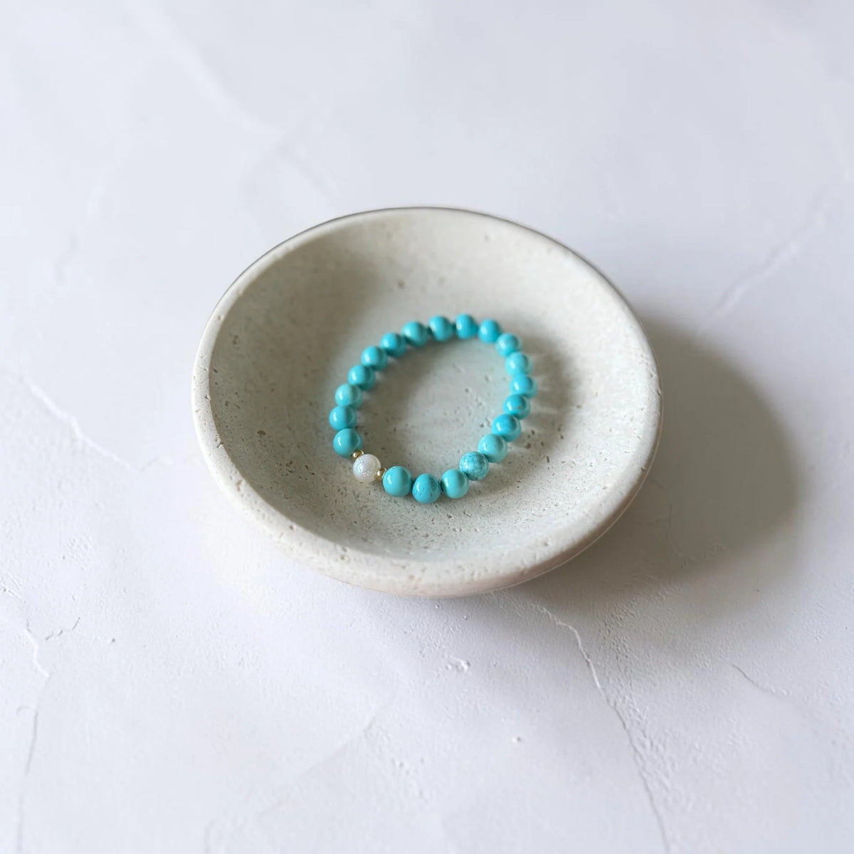 A turquoise beaded stretch bracelet with a single white pearl accent, displayed inside a small speckled ceramic dish on a light textured surface.