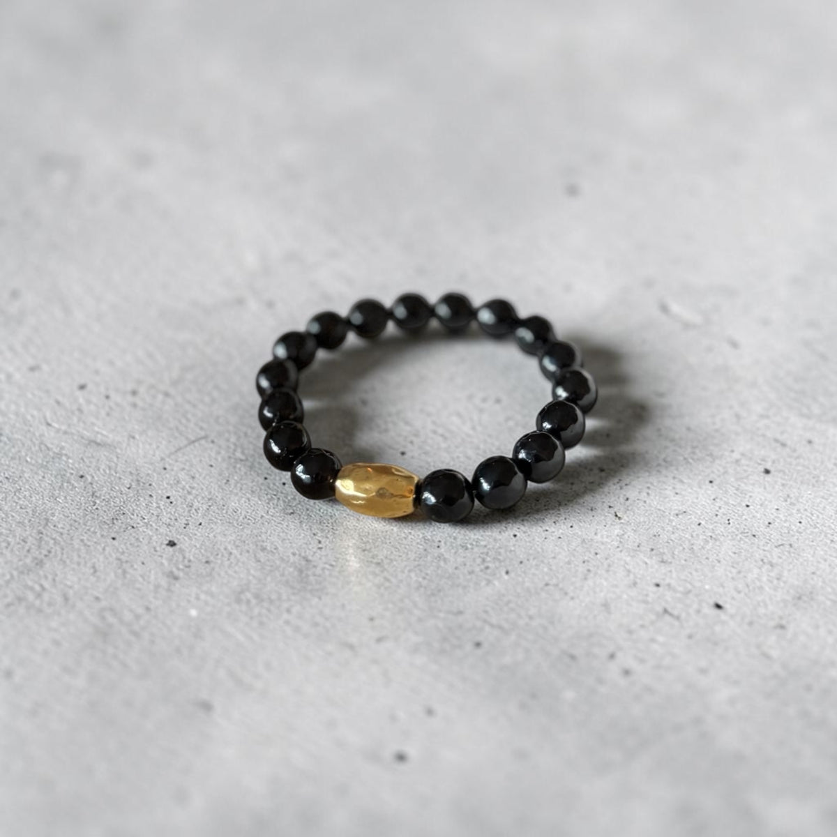 A beaded stretch bracelet featuring glossy black round beads and a single faceted gold accent bead, resting on a textured gray concrete surface.