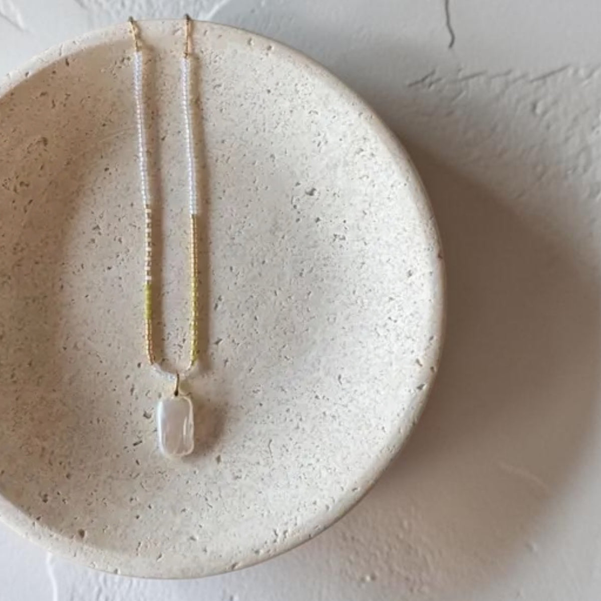 A delicate necklace with a small white irregular pendant, displayed on a textured beige ceramic dish against a light neutral background.