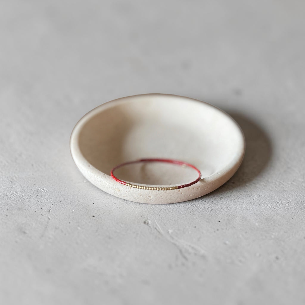 A small round ceramic dish or shallow bowl in warm off-white, with a thin red ring inside and a tiny gold line along the inner edge, photographed on a light gray textured surface.
