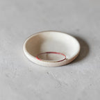 A small round ceramic dish or shallow bowl in warm off-white, with a thin red ring inside and a tiny gold line along the inner edge, photographed on a light gray textured surface.