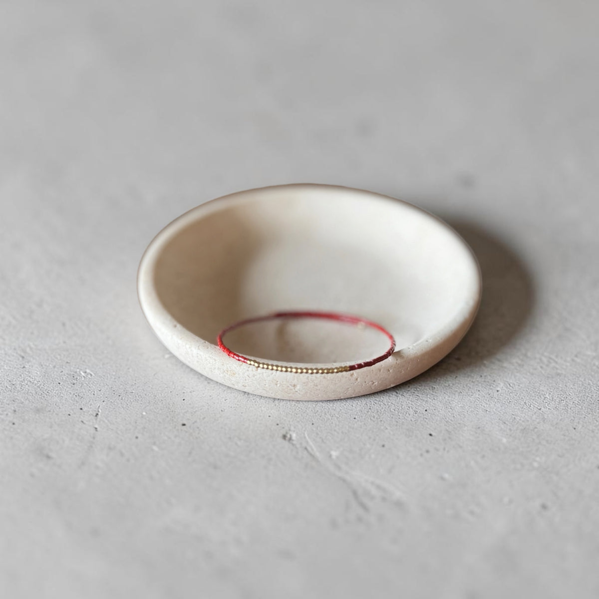 A small round ceramic dish or shallow bowl in warm off-white, with a thin red ring inside and a tiny gold line along the inner edge, photographed on a light gray textured surface.