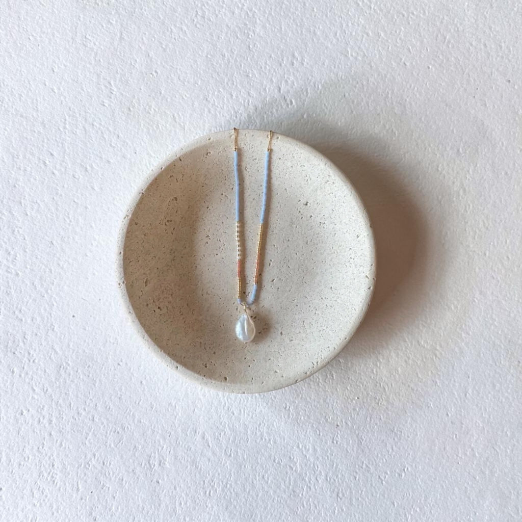 A delicate necklace with a white pearl pendant and thin gold and light blue chains, displayed on a small round beige ceramic dish against a textured white background.
