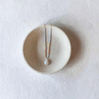 A delicate necklace with a white pearl pendant and thin gold and light blue chains, displayed on a small round beige ceramic dish against a textured white background.