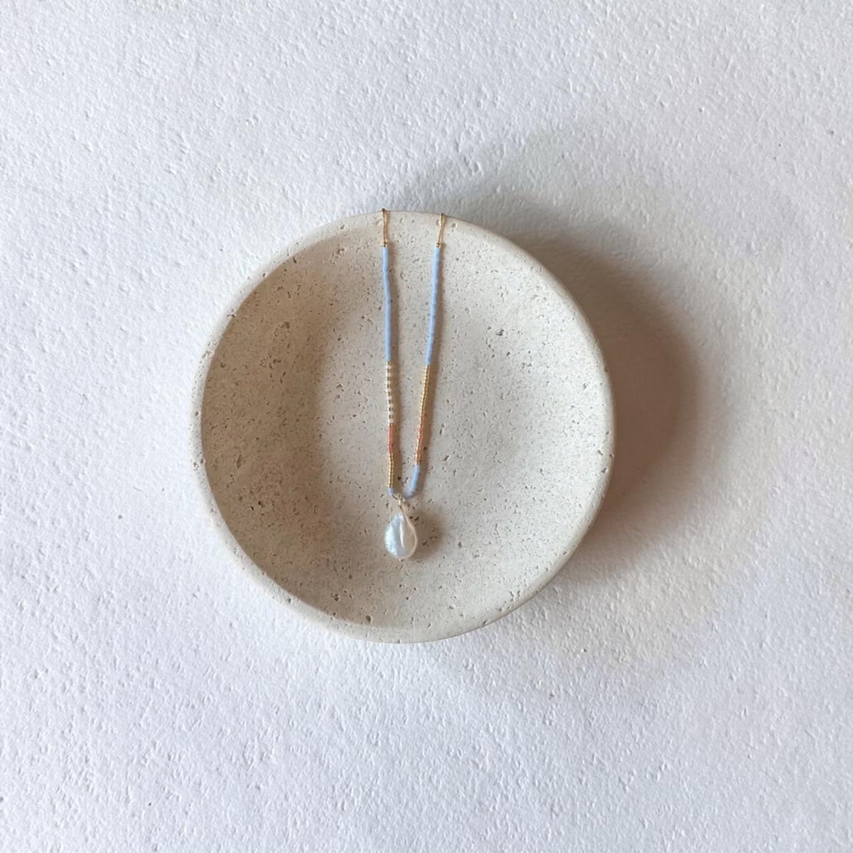 A delicate necklace with a white pearl pendant and thin gold and light blue chains, displayed on a small round beige ceramic dish against a textured white background.