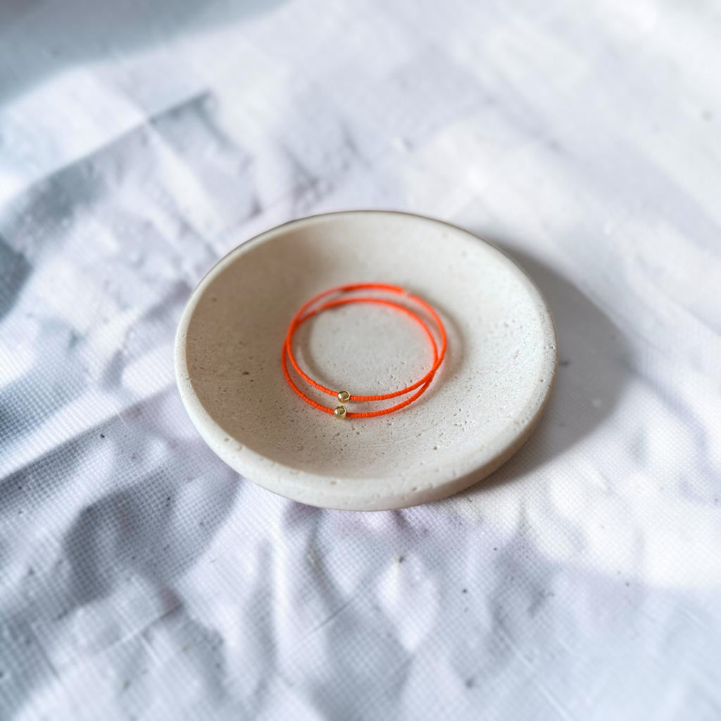 Two thin orange wire bracelets with small gold bead details resting inside a small round beige ceramic dish on a white textured fabric background.