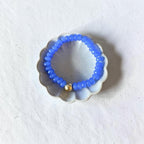 A beaded stretch bracelet in bright blue with a single small gold bead accent, displayed on a scalloped white dish against a textured white background.