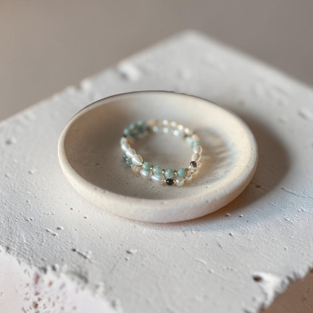 A delicate beaded bracelet with small white pearls and pale mint-green beads, displayed on a small round ceramic dish atop a textured white concrete block.