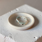 A delicate beaded bracelet with small white pearls and pale mint-green beads, displayed on a small round ceramic dish atop a textured white concrete block.
