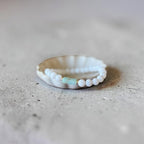 A delicate beaded bracelet with white round beads and a small mint green rectangular accent, displayed on a white scalloped dish over a textured gray concrete surface.