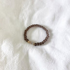 A beaded stretch bracelet with dark brown translucent round beads and a small light beige accent bead, displayed on a textured white fabric background.