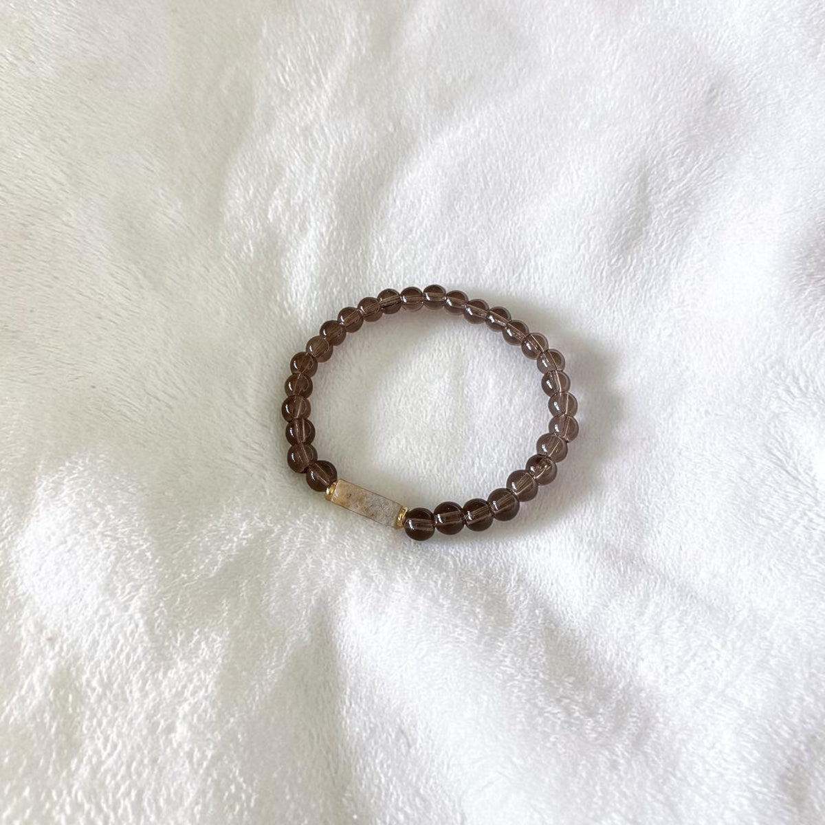 A beaded stretch bracelet with dark brown translucent round beads and a small light beige accent bead, displayed on a textured white fabric background.