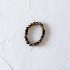 A beaded stretch bracelet made of brown and black tiger's eye beads with a single dark blue-gray iridescent bead, displayed on a textured white surface.