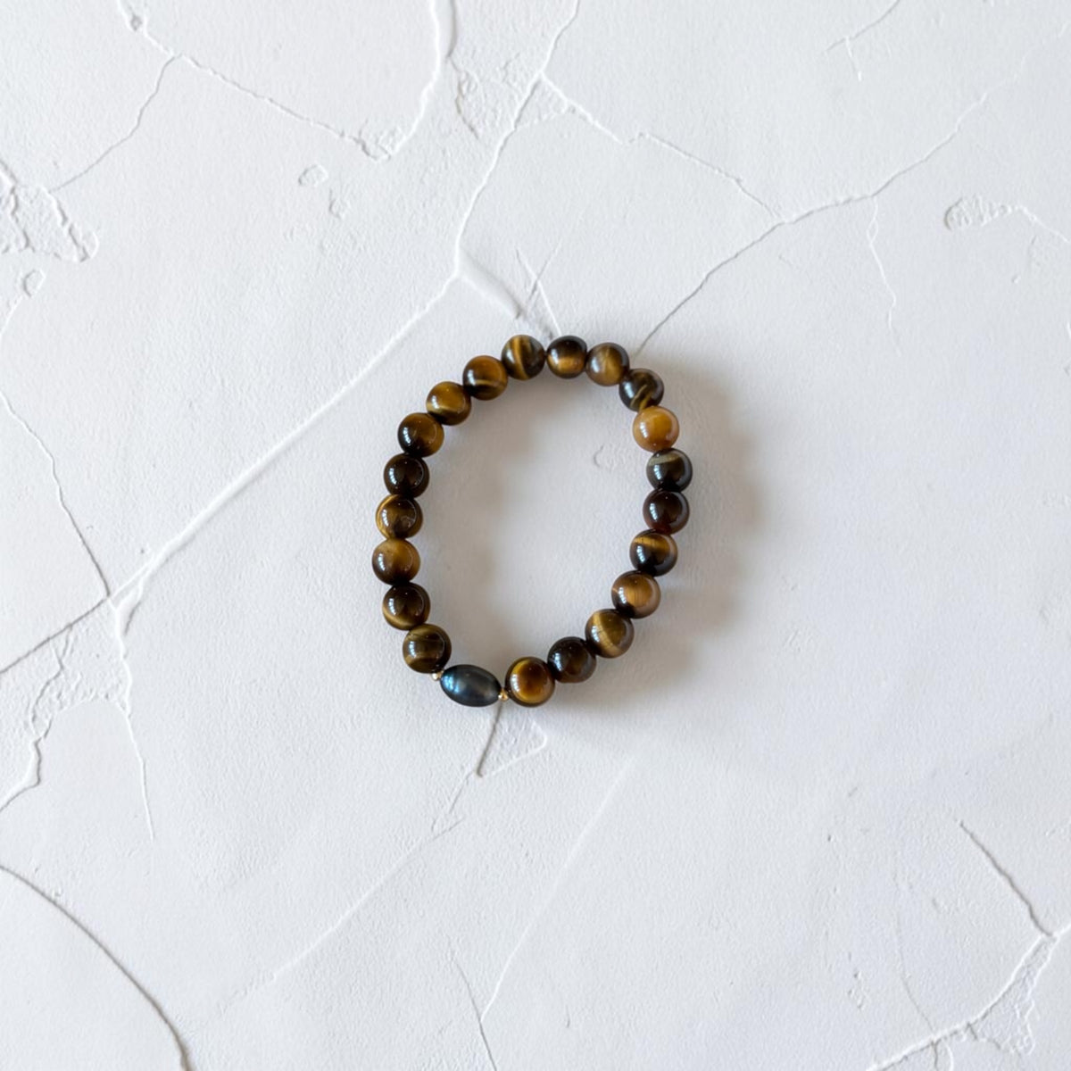 A beaded stretch bracelet made of brown and black tiger's eye beads with a single dark blue-gray iridescent bead, displayed on a textured white surface.