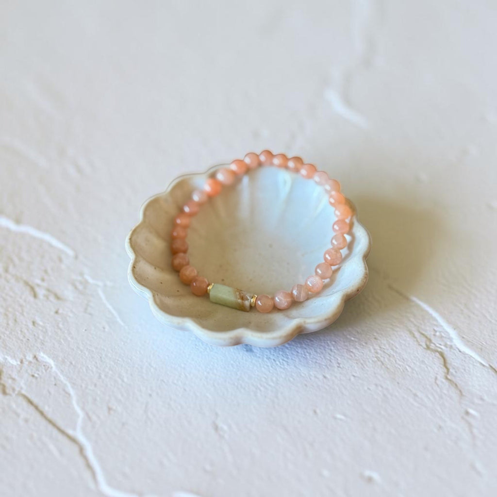 A beaded stretch bracelet in peachy pink and light green, displayed inside a small scalloped ceramic dish on a textured off-white surface.