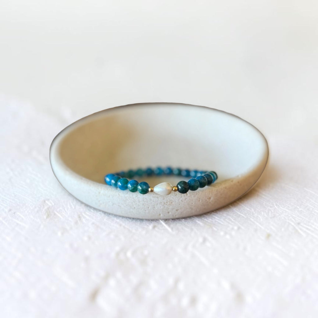 A beaded stretch bracelet with teal blue beads and a small white pearl accent, displayed inside a small, shallow, off-white ceramic dish on a textured white surface.