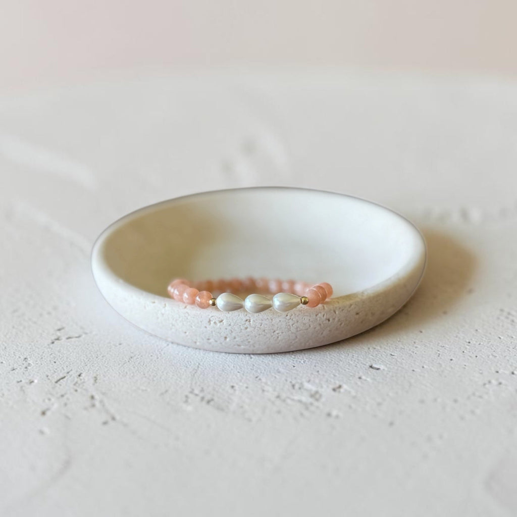 A delicate beaded bracelet with pink and white beads resting inside a small, round, textured ceramic dish on a light, neutral surface.