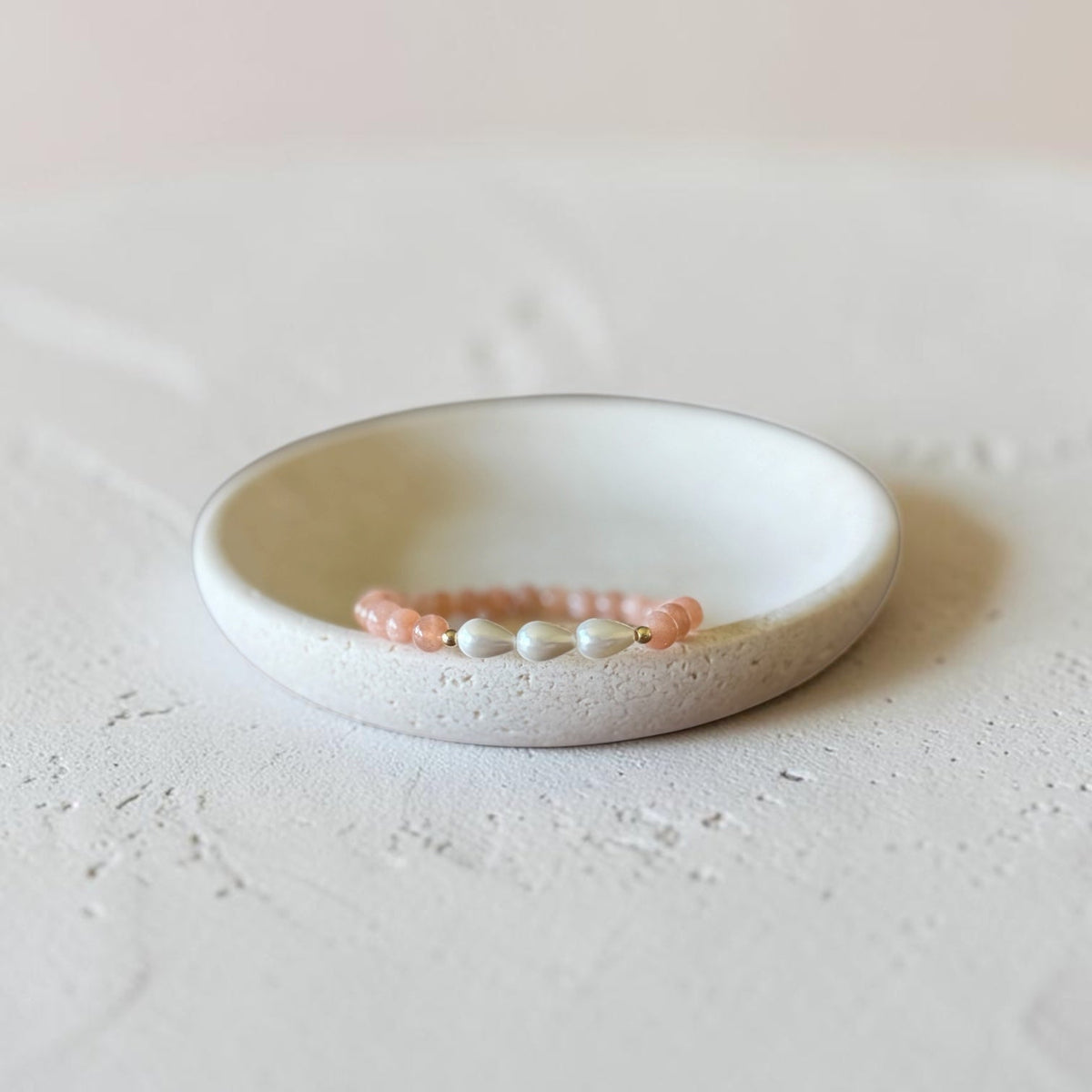 A delicate beaded bracelet with pink and white beads resting inside a small, round, textured ceramic dish on a light, neutral surface.