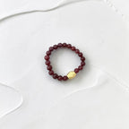 A beaded stretch bracelet featuring dark red faceted beads and a single gold-toned geometric accent bead, displayed on a textured white surface.