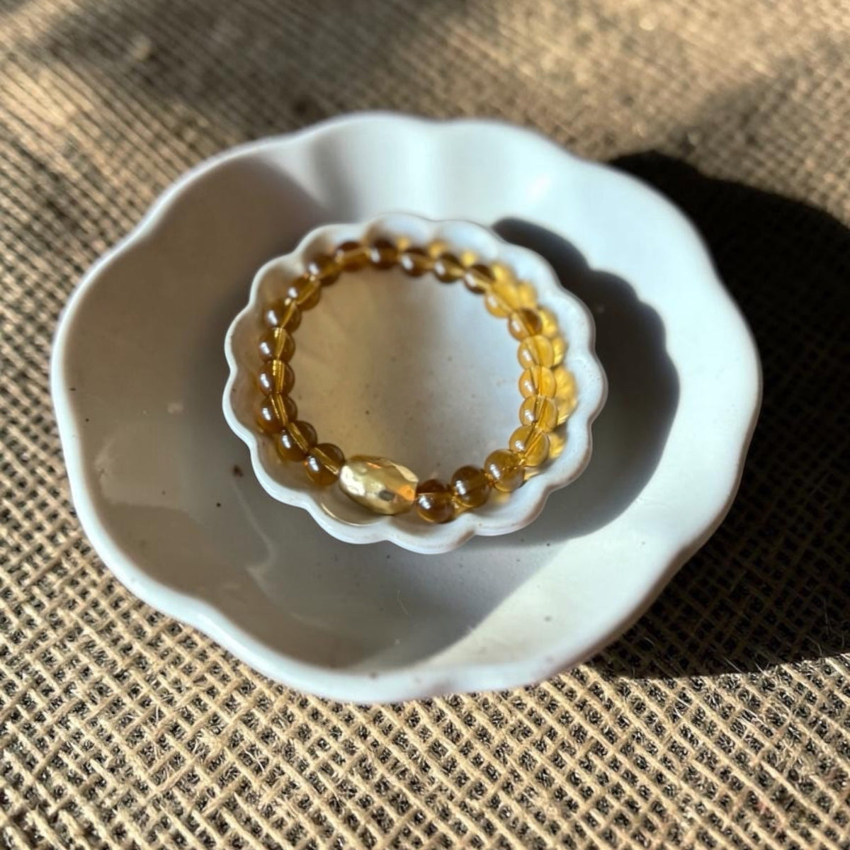 A beaded stretch bracelet made of translucent amber-yellow beads with a central faceted yellow stone, displayed inside a small white ceramic dish on a textured tan woven fabric background.