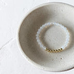 A beaded stretch bracelet with translucent white beads and a small gold bead accent, displayed inside a speckled ceramic dish on a white textured surface.