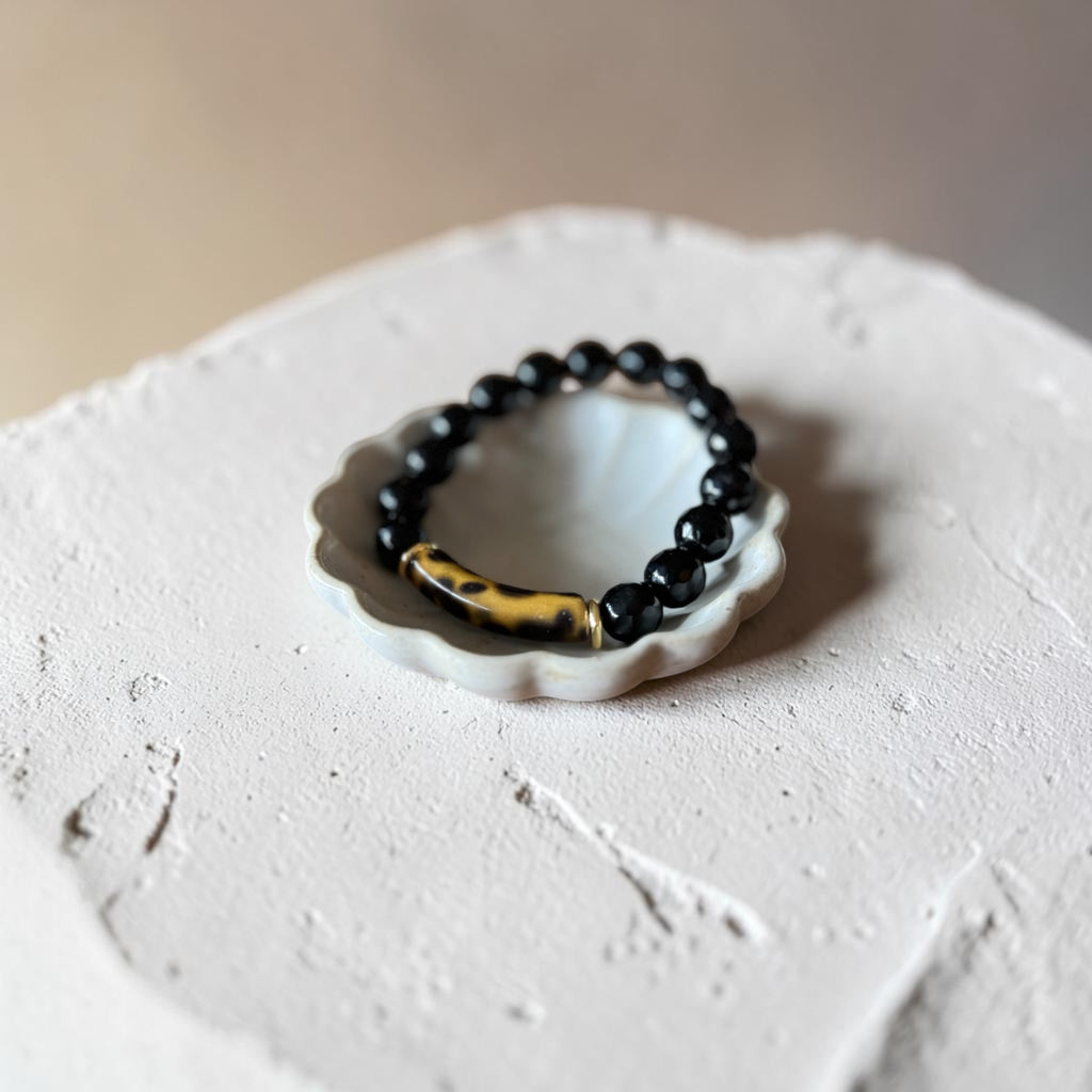 A beaded stretch bracelet with glossy black round beads and a central gold-toned bar with black spots, displayed on a small white scalloped dish over a textured light surface.