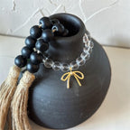 A beaded necklace with matte black round beads, a strand of clear faceted beads, and a small gold bow charm, displayed on a matte black ceramic vase against a light textured wall.