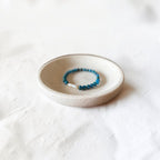 A beaded stretch bracelet with blue beads and a silver-toned clasp, displayed on a small round ceramic dish over a textured white fabric background.