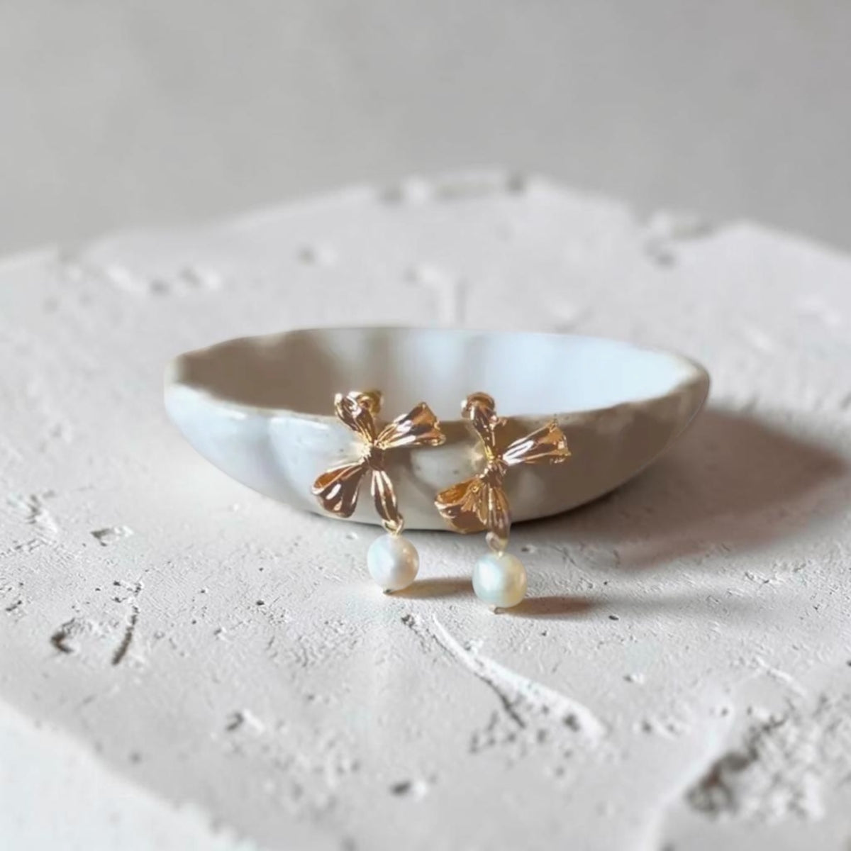 A pair of gold bow drop earrings with white pearl beads, displayed on a small white ceramic dish against a textured light gray background.