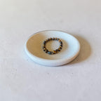 A beaded stretch bracelet with dark brown and metallic gold beads resting on a small, round, light gray ceramic dish against a neutral light gray background.