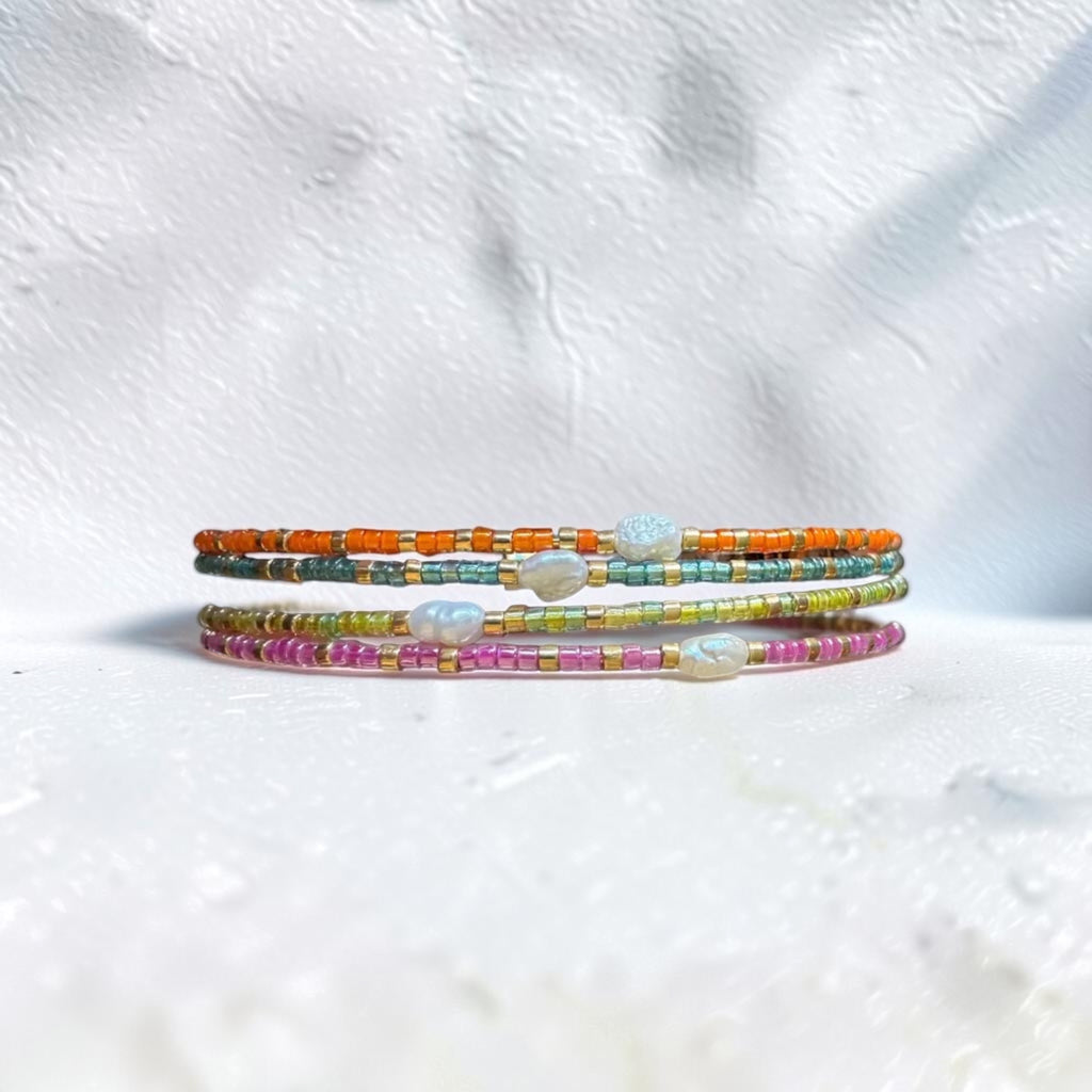 Three thin beaded bracelets stacked together on a textured white surface. The bracelets feature small multicolored beads in orange, teal, yellow-green, and pink, with small white shell-like accents.