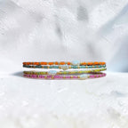 Three thin beaded bracelets stacked together on a textured white surface. The bracelets feature small multicolored beads in orange, teal, yellow-green, and pink, with small white shell-like accents.