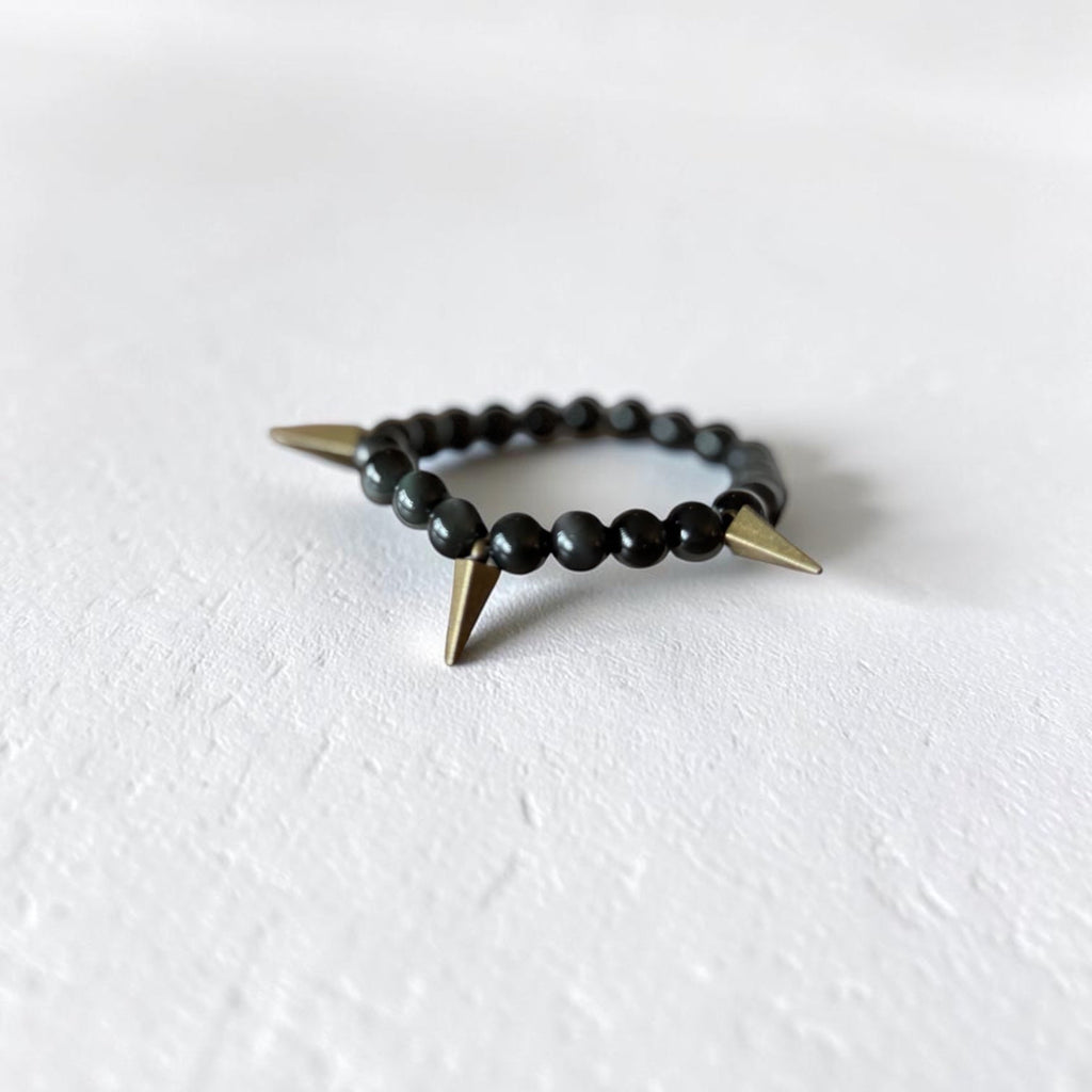 A beaded stretch bracelet featuring matte black round beads and two sharp, metallic gold spikes, resting on a textured white surface.