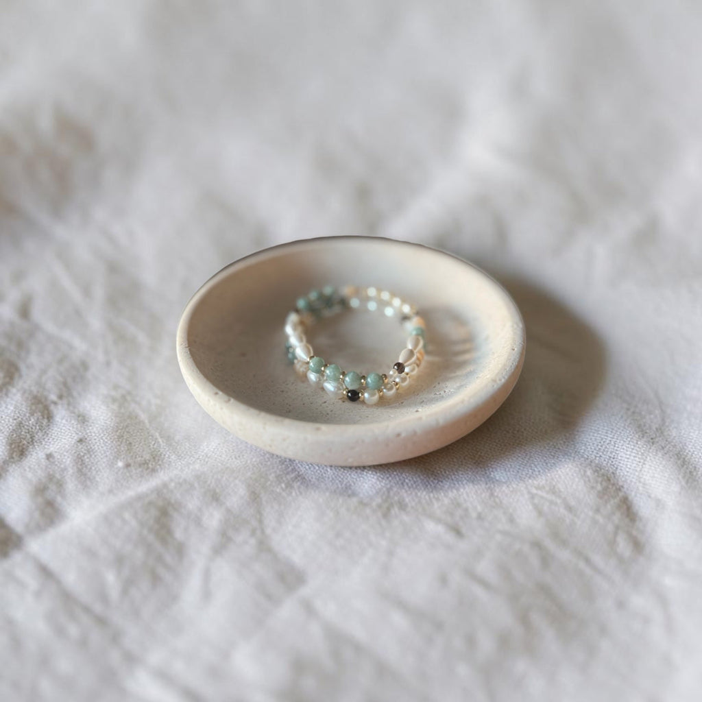 A delicate beaded bracelet with small turquoise, silver, and dark beads resting on a small round ceramic dish over a textured white fabric background.