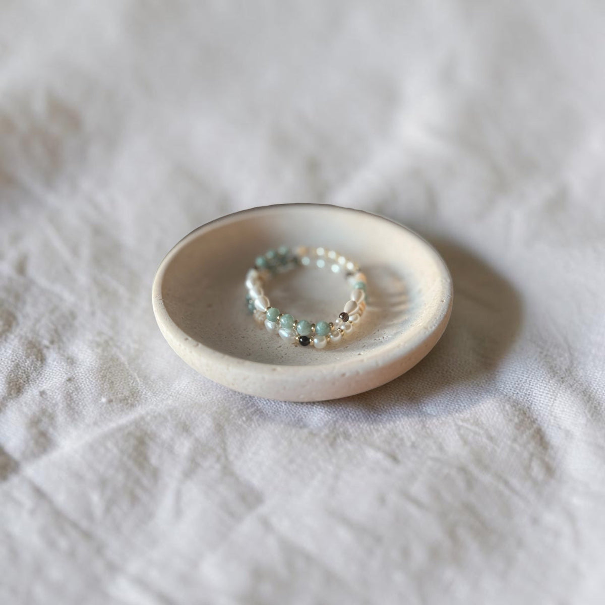 A delicate beaded bracelet with small turquoise, silver, and dark beads resting on a small round ceramic dish over a textured white fabric background.