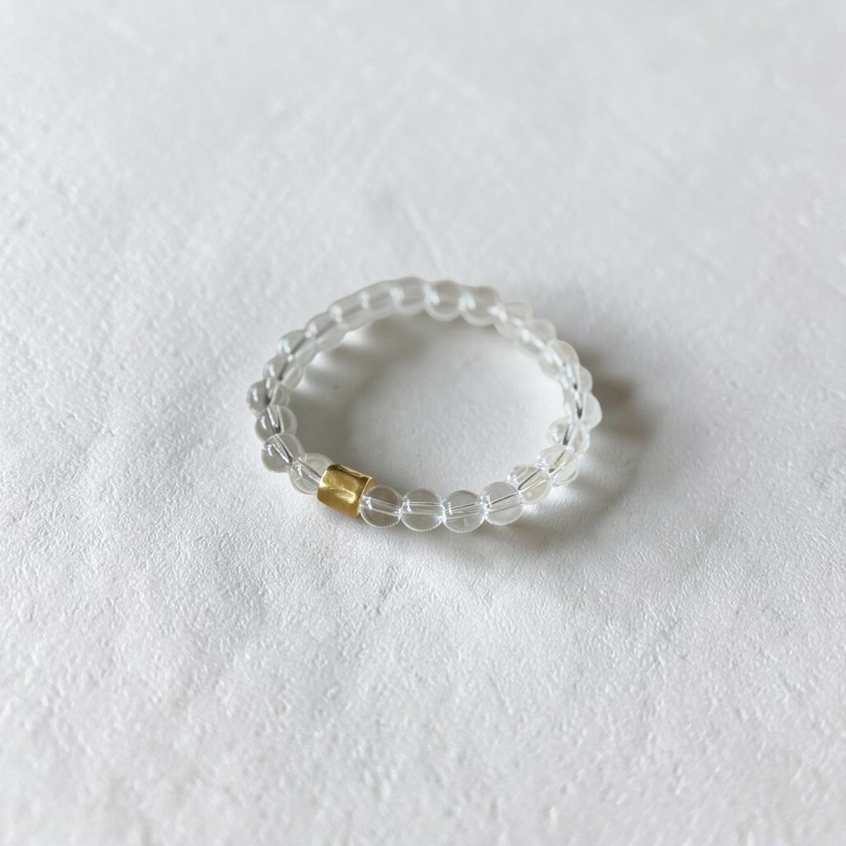 A clear beaded stretch bracelet with a small gold accent bead, photographed on a textured white surface.