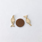 Two small gold fish charms displayed on a white textured surface, with a brown coin placed between them for scale.