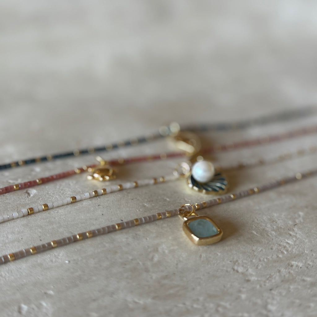 A close-up, shallow depth-of-field shot of several delicate beaded strands and pendants arranged on a textured light beige surface. The jewelry includes gold-toned chains with small gold beads, a pearl pendant, a dark fan-shaped charm, and a square pendant with a pale blue-gray stone in a gold bezel.